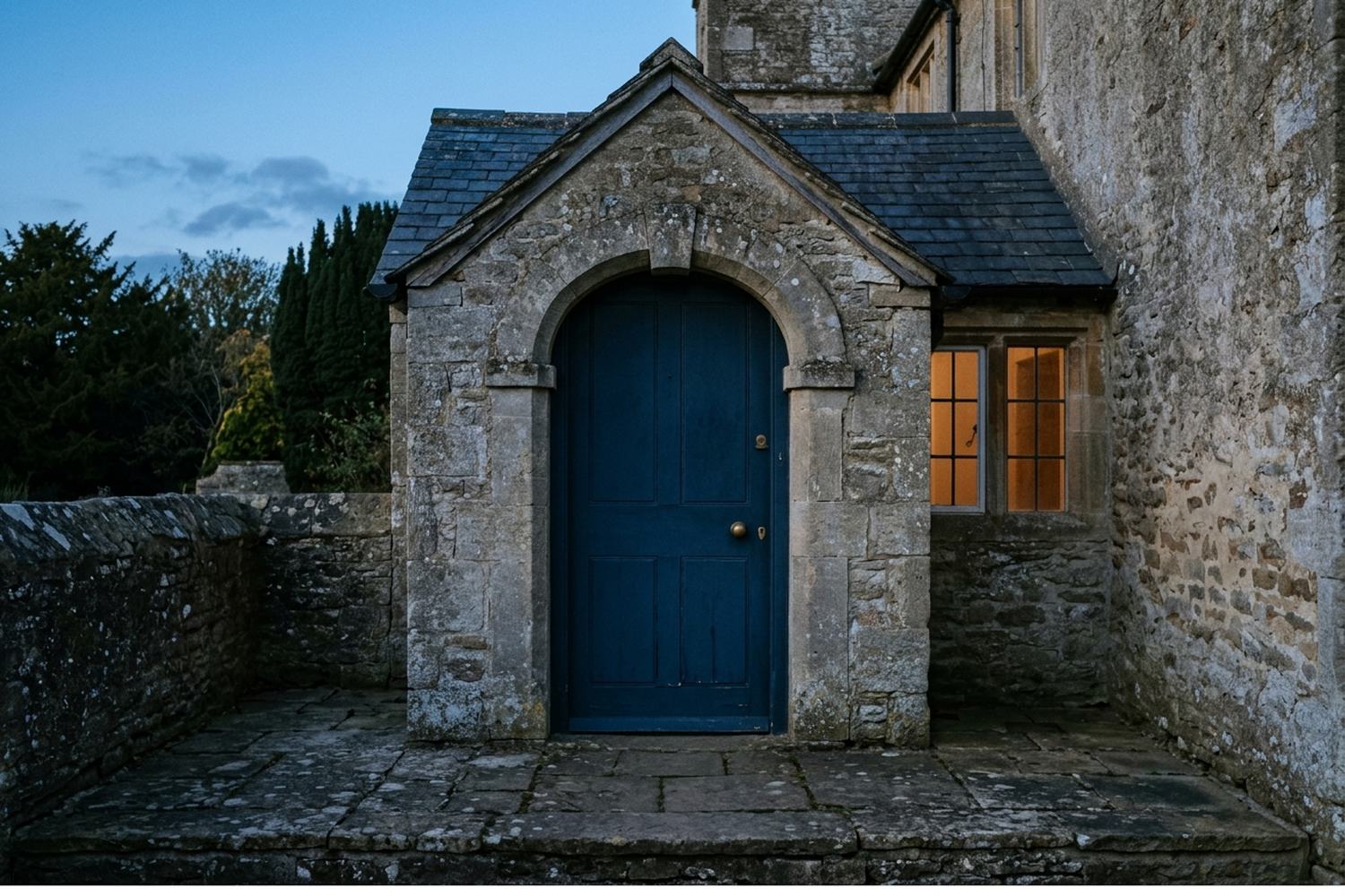 A plain door under a stone porch at dusk with one warm light beside it.
