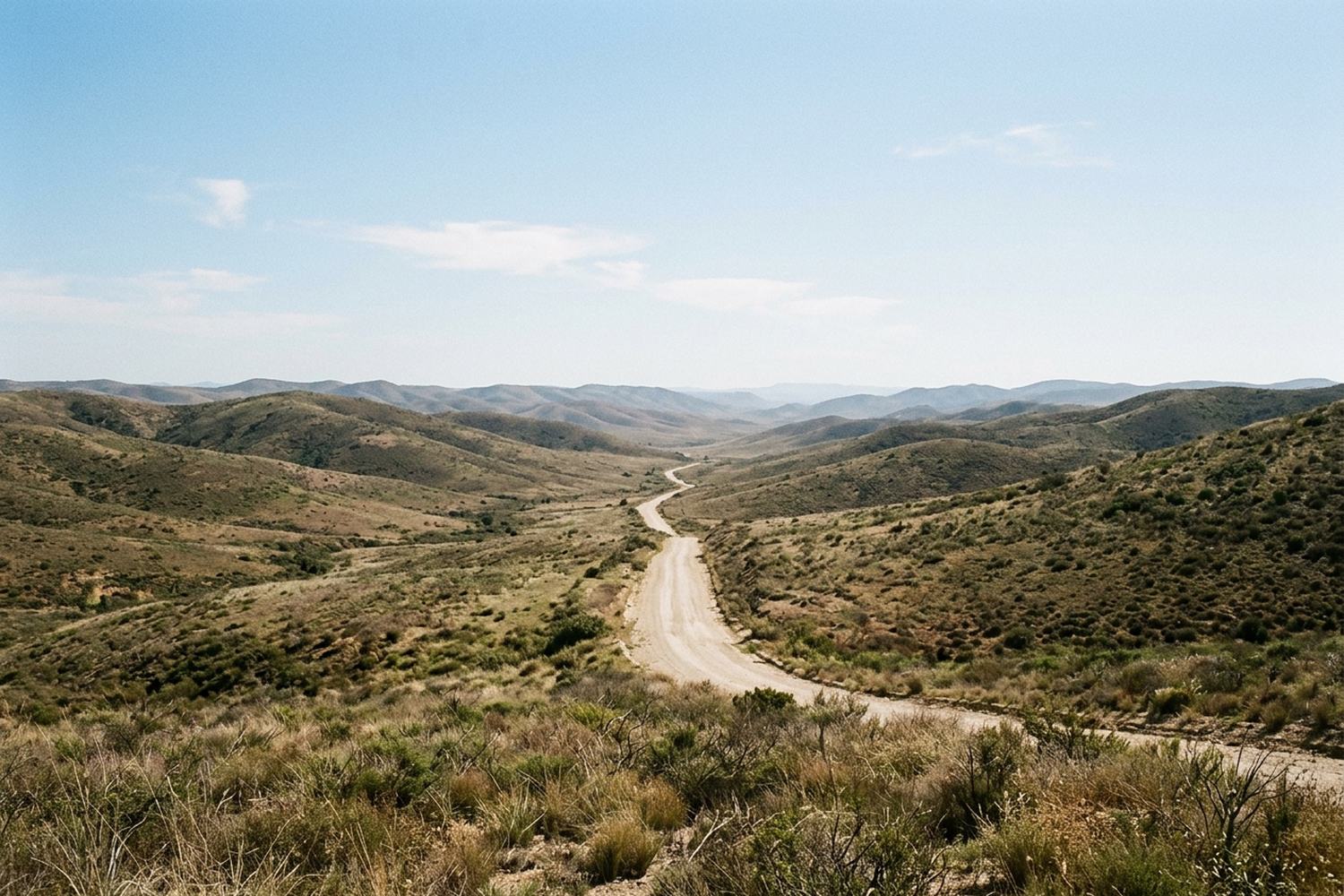 Range behind range of low hills at midday with a pale road running between them.