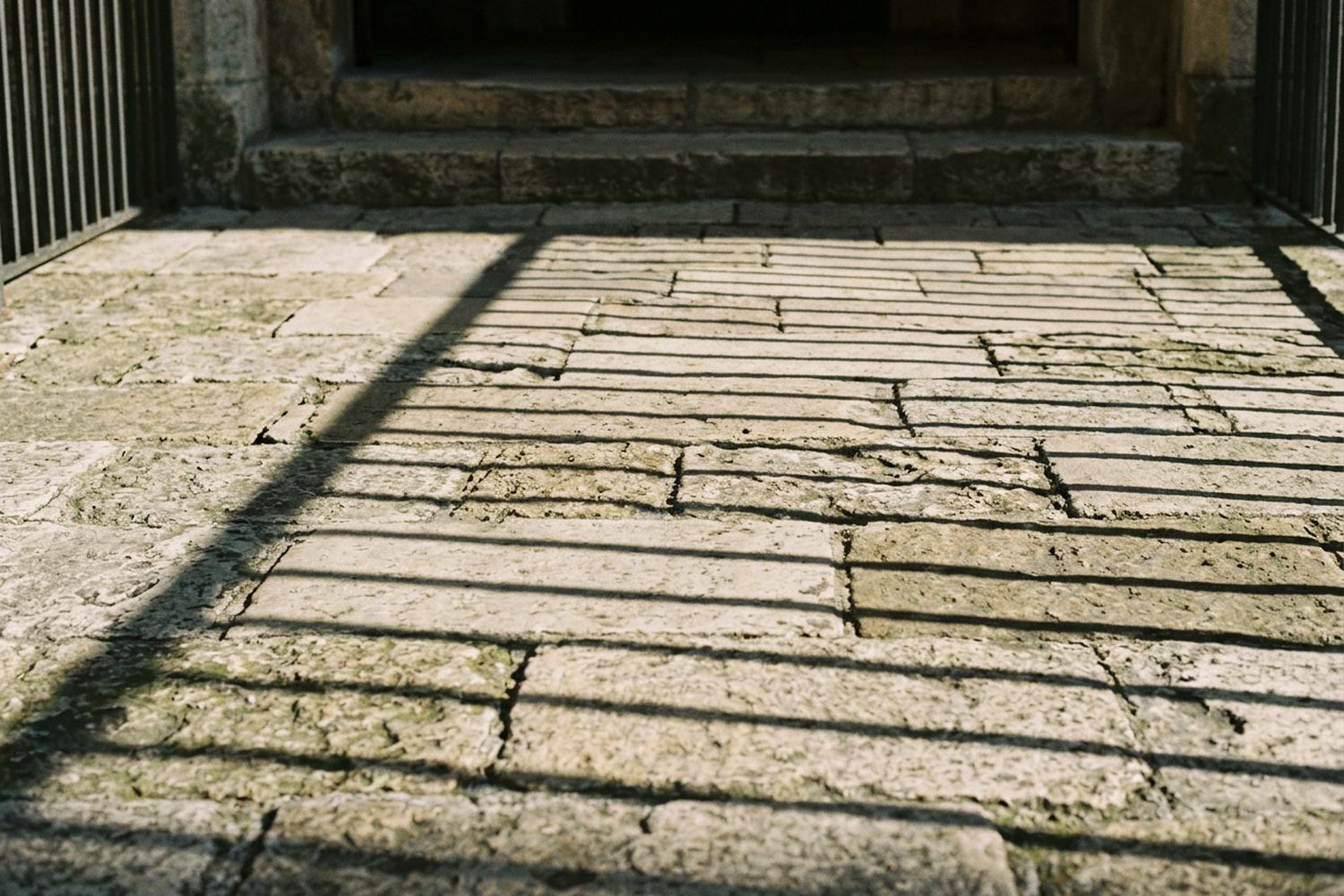 Long early shadows of an iron railing thrown in stripes across a worn stone floor.